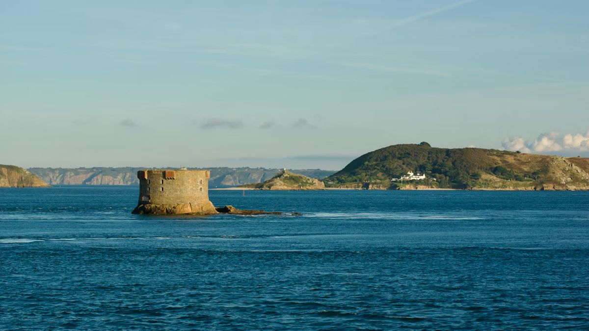 A granite tower on a rock in the sea off Guernsey, Channel Islands, with small green islands in front and Sark on the horizon.