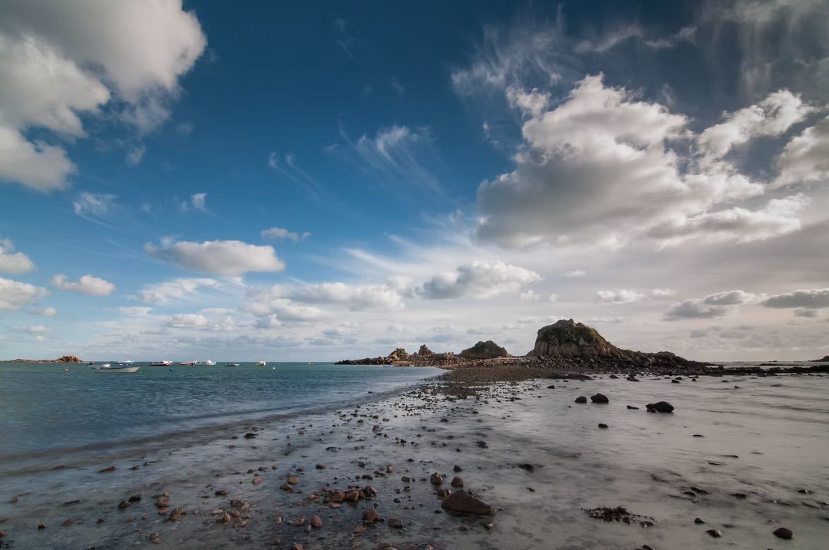 Rocks and wet sand on the shore at Le Hocq, St Clement, Jersey, Channel Islands, under a windswept sky, with boats moored offshore.
